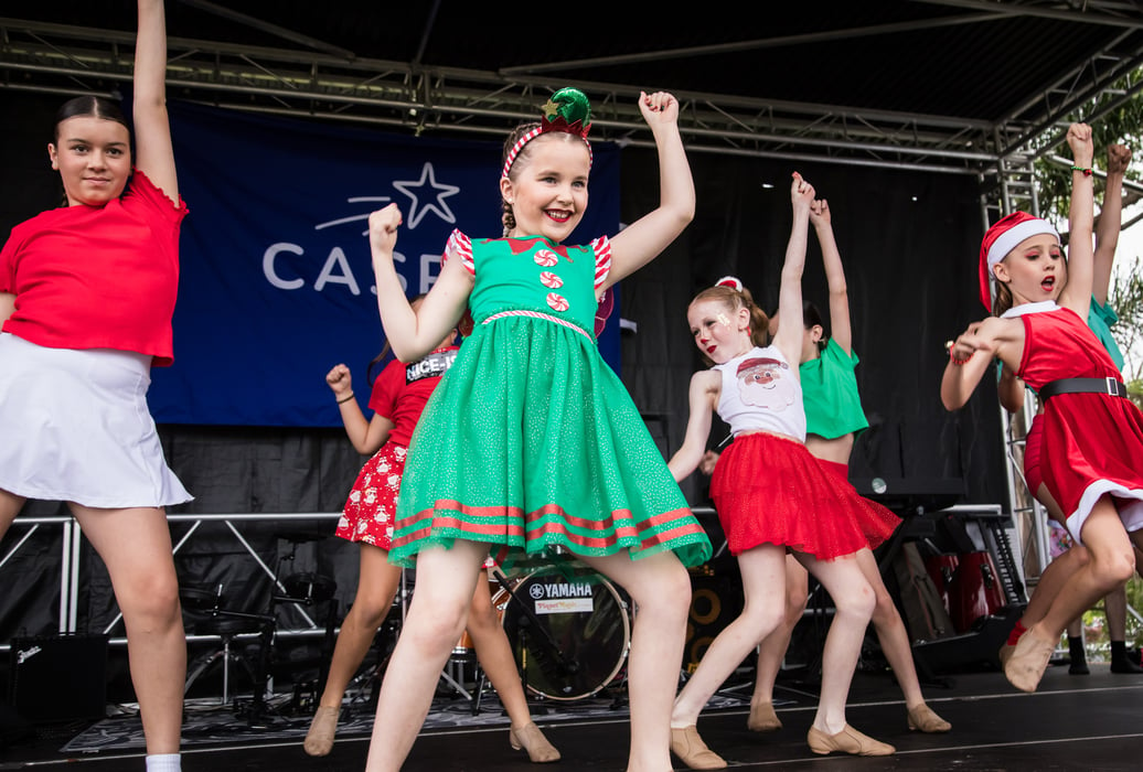 A group of girl's dressed in festive green and red costumes dance energetically on a stage in front of a blue banner with a shooting star that reads CASPA 