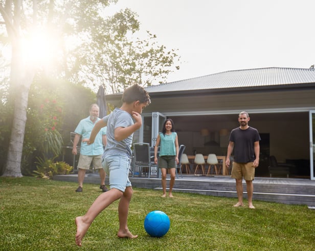 A young boy playing soccer in the backyard while three smiling adults watch and cheer.