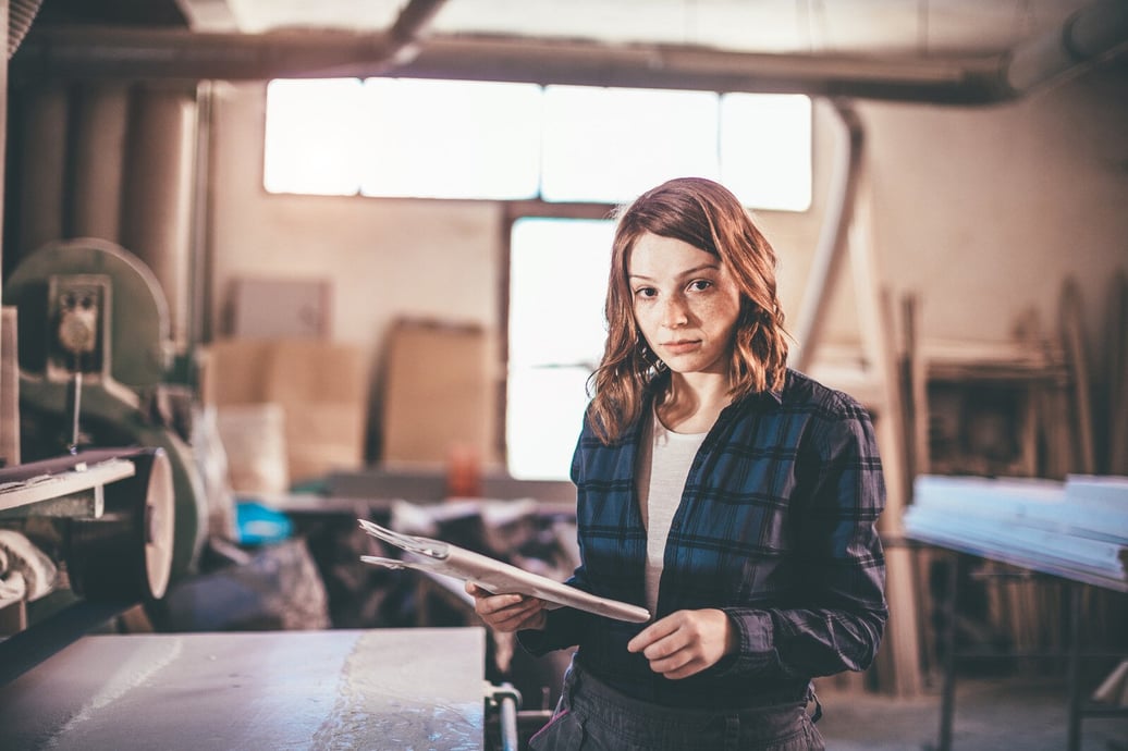 A young person stands in a well lit workshop, reviewing paperwork. She is wearing a flannel shirt and work pants.