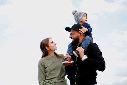 A couple walk happily together outside. A young child sits on the man's shoulders.