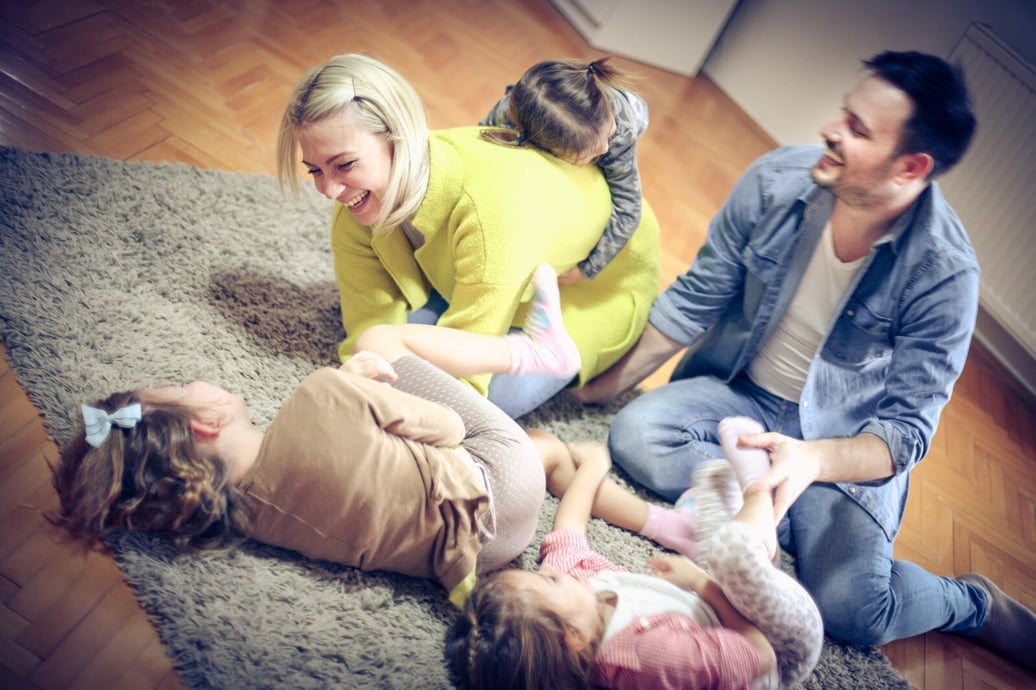 A mum and dad laugh and play with their three daughters on the floor in a living room setting.