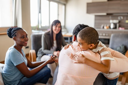 A family attends a therapy session, the father and son embracing. The setting is comfortable and light.