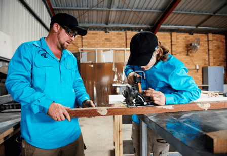 A young person works with his mentor on carpentry in an open, light workshop. They both wear bright blue work shirts and safety gear.