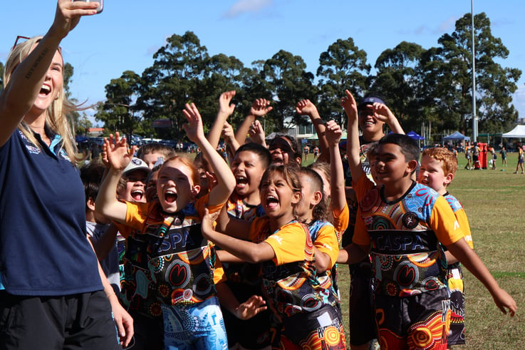 A group of diverse children in Indigenous printed shirts happily posing for a photo with a CASPA employee in an outdoor setting