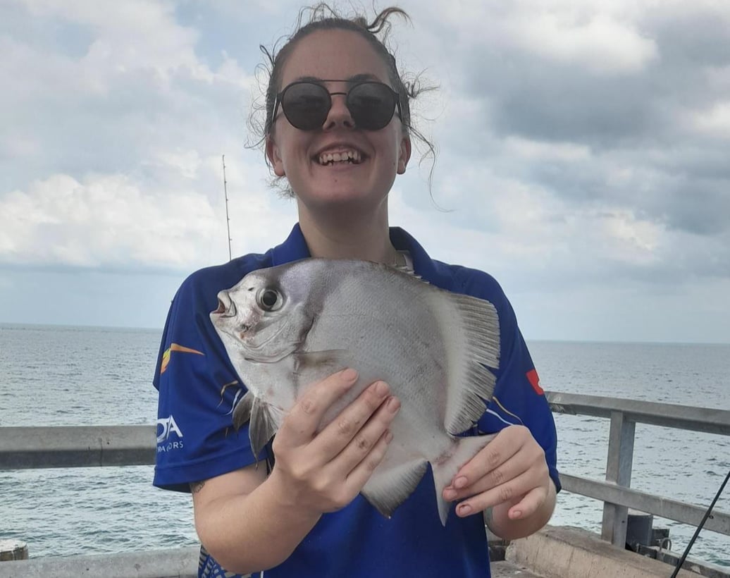 A smiling young person in blue stands on a jetty holding a fish they caught.