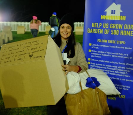 A white woman with long dark hair stands in front of the CEO Sleepout sign. She is wearing warm clothes and holding a large back and a cardboard box with her message of hope written on it.