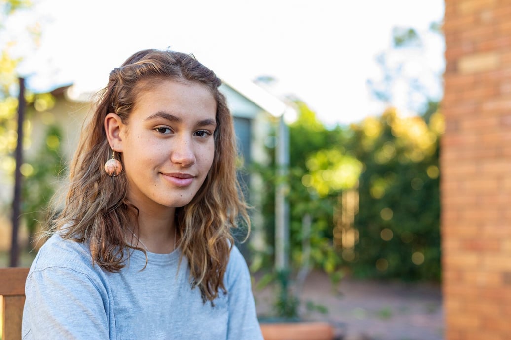 A young, Indigenous girl with a half smile on her face looks at the camera. She is standing in a bright, open garden.