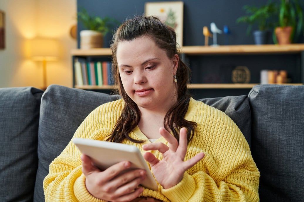 A young woman with down syndrome, wearing a yellow sweater, sits comfortably using a tablet in a warm, lounge room setting