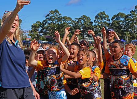 Excited young people crowd together with hands in the air for a photo, wearing CASPA football jerseys with Aboriginal designs on them.