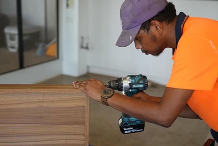 A young Indigenous man dressed in high-res clothing works with a drill on some cabinetry