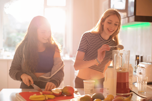 Two teenage girls make food together in sunlit kitchen