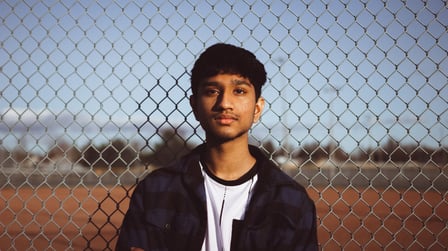 A teenage boy with tawny skin and dark, short hair, stares straight at the camera. He leans against a chainmail fence.