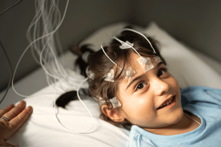 A young girl with olive skin and dark hair calmly looks past the camera. She lies in a bed with neuro-feedback sensors attached to her head. Part of an adult's hand sits at the edge of the frame, indicating she isn't alone