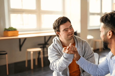 A smiling white man with down syndrome is warmly greeting a another man who is mostly out of view.