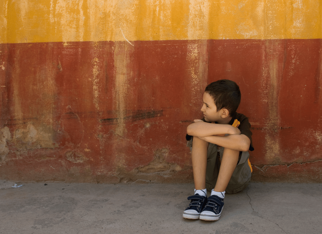 Young white boy sits alone outside on the concrete, holding himself and looking away from the camera