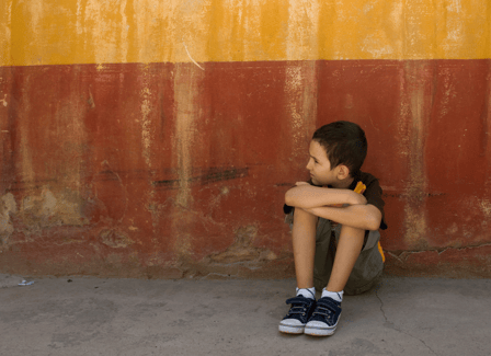 Young white boy sits alone outside on the concrete, holding himself and looking away from the camera