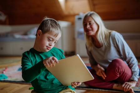 A boy with down syndrome and light skin is engaged in an activity. An adult woman with pale skin and blonde hair sits behind him out of focus, observing his actions.