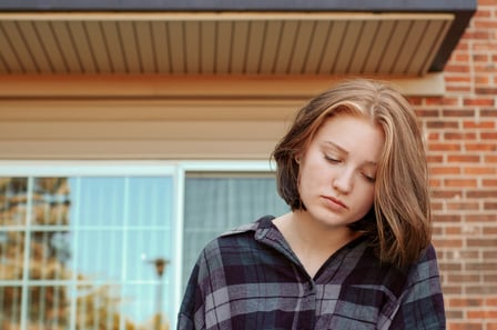 A young woman with pale skin and straight hair sits alone in front of a brick house. She looks sad, her gaze towards the ground