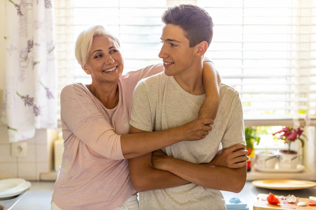 A mother and teenage son, embracing and smiling, stand in a kitchen filled with natural light.